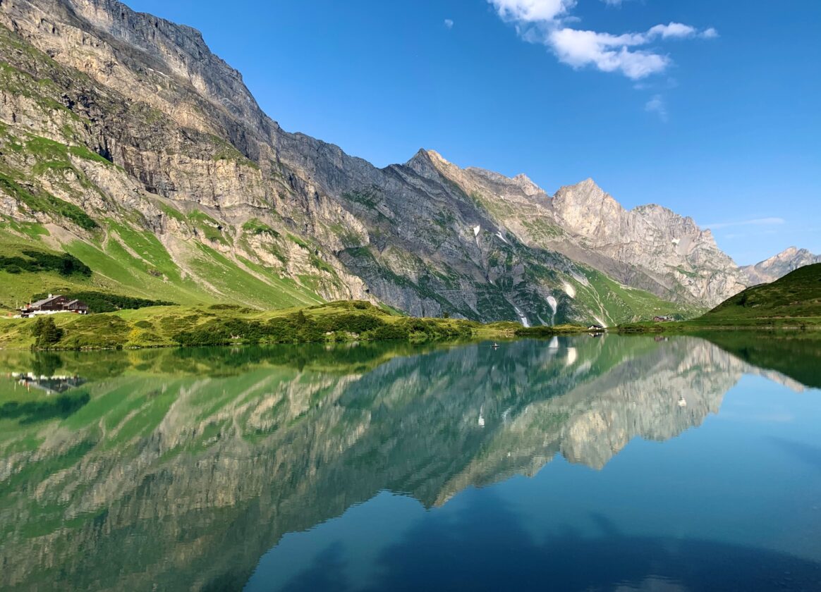 Berglandschaft mit See, grünen Hängen und felsigen Gipfeln unter blauem Himmel. Die Berge spiegeln sich im ruhigen Wasser.
