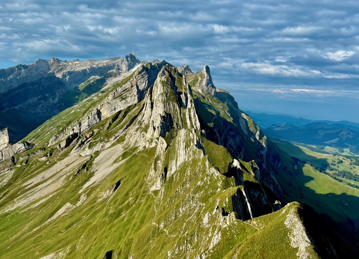 Markanter, grasbewachsener Berggrat mit steilen Felsformationen. Im Hintergrund erstrecken sich weitere Bergketten und Täler unter einem dicht bewölkten Himmel.