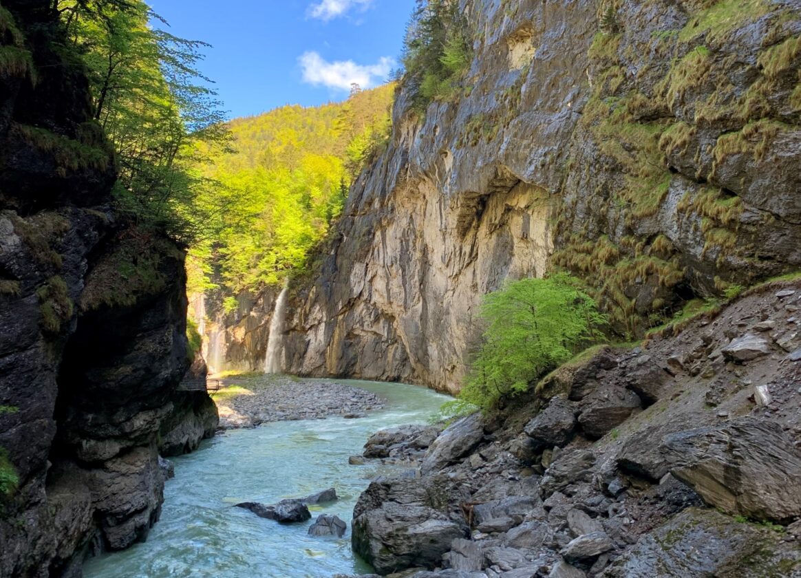 Türkisfarbener Fluss in einer engen, felsigen Schlucht. Steile, teils moosbewachsene Felswände und grüne Laubbäume umgeben das Gewässer; im Hintergrund fällt ein kleiner Wasserfall in die Schlucht.