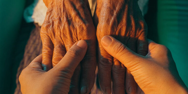 Zwei jüngere Hände halten behutsam die gefalteten, stark gefurchten Hände einer älteren Person. Warmes Licht hebt die Hände vor einem dunkelgrünen Hintergrund hervor.